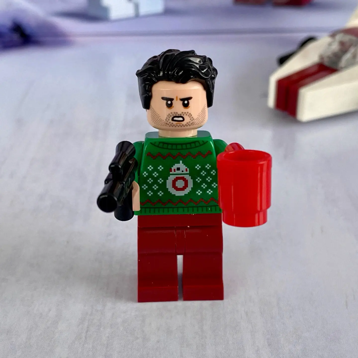 LEGO minifigure Poe Daemeron wearing a green knitted holiday sweater with BB-8 pattern. Poe holds a red coffee cup in one hand and a blaster in the other.