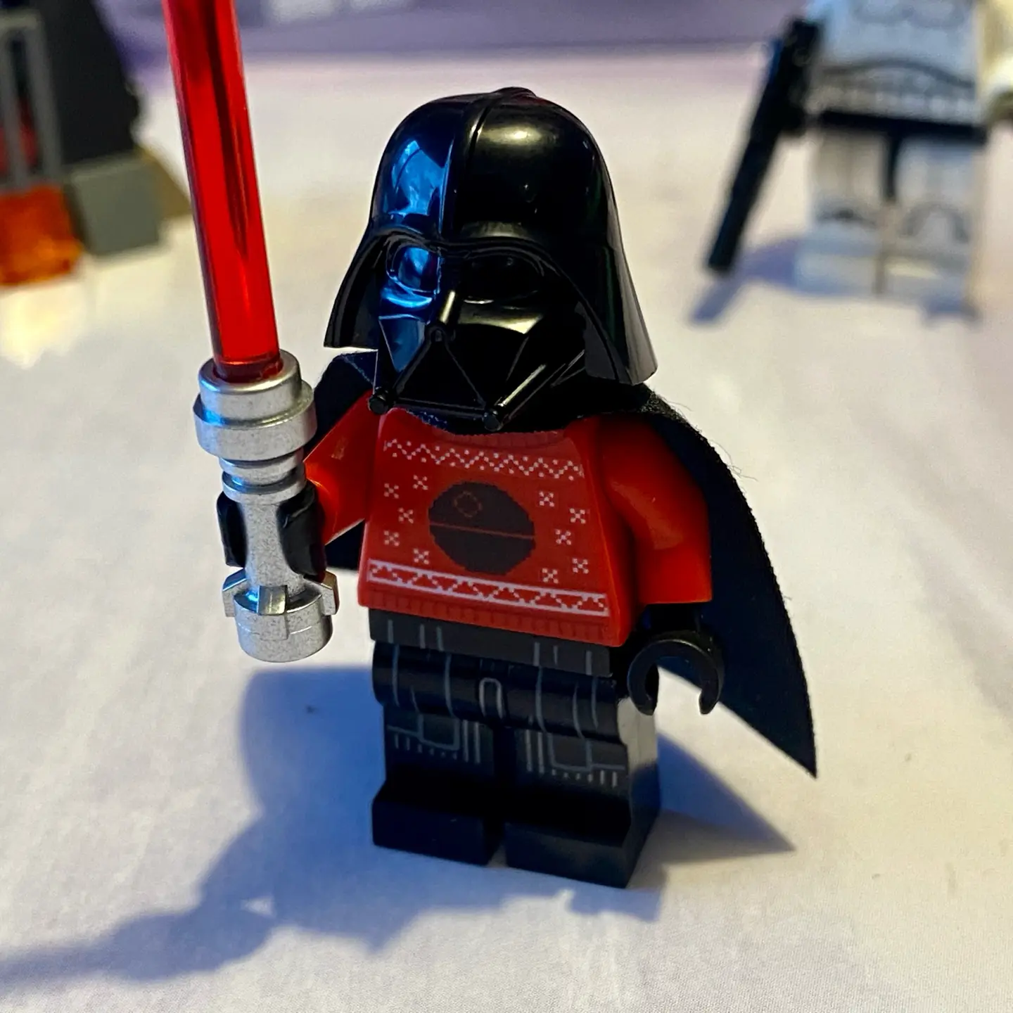 LEGO minifigure Darth Vader wearing a red knitted holiday sweater with Death Star pattern. Darth Vader holds his red light saber in one hand.