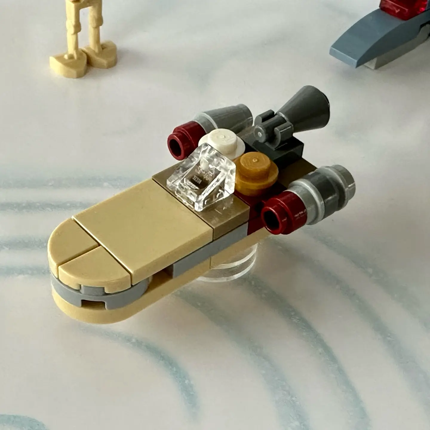 LEGO micro-scale build of Luke's landspeeder in tan with two quarter-circle tiles used to shape the hood. White and pearl gold round studs represent Luke and C-3PO. The left engine has a different color and shape because its cowling had been removed.