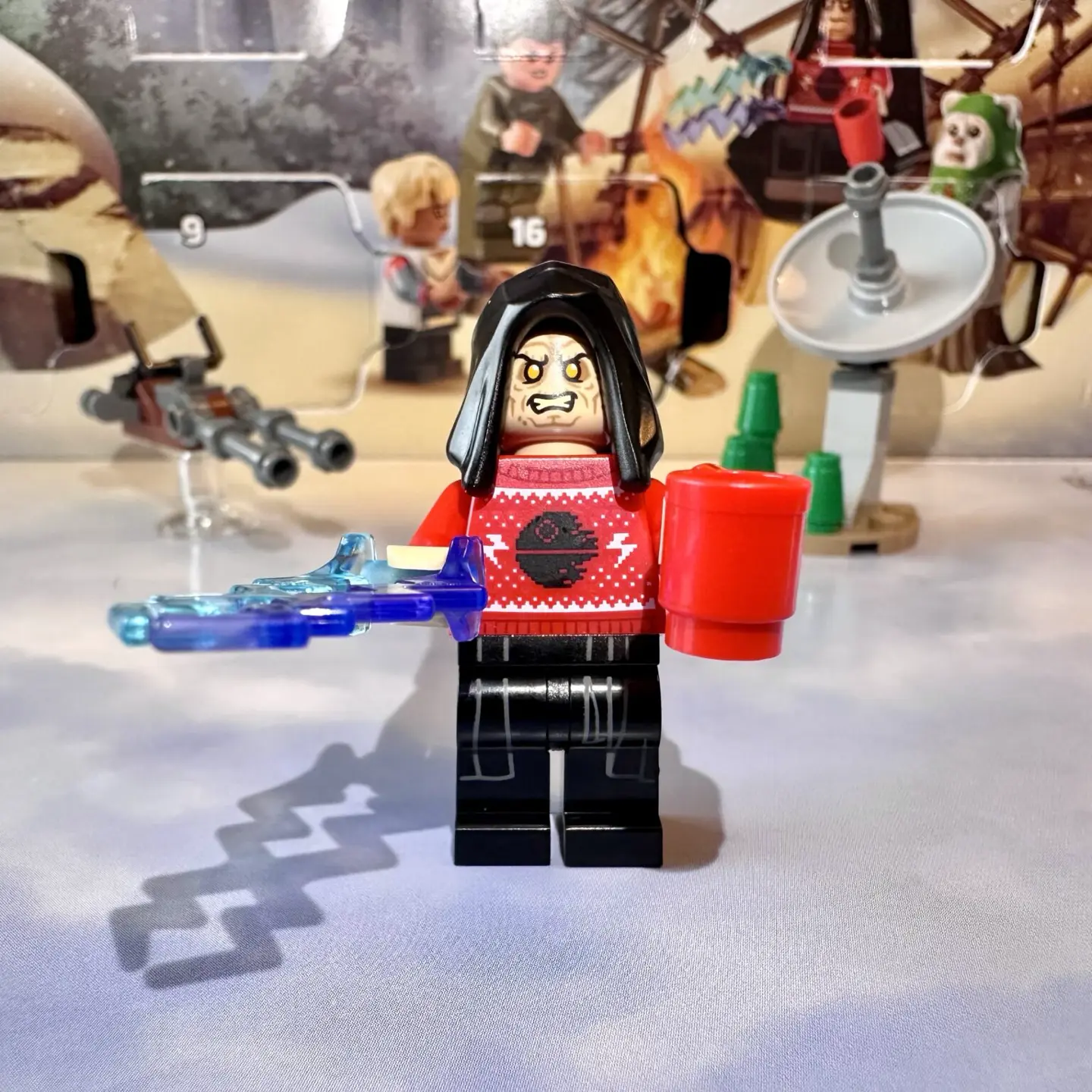 LEGO minifigure Emperor Palpatine wearing a red knitted holiday sweater with Death Star pattern. Palpatine holds a red coffee cup in one hand and blasts energy bolts with the other.