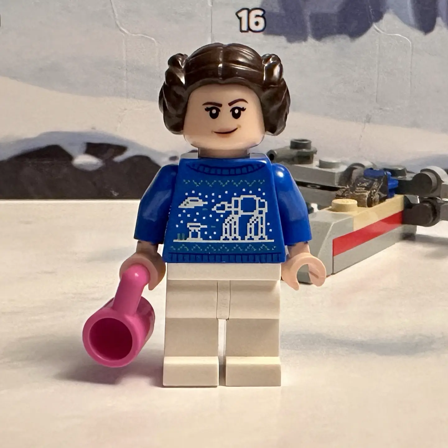 Princess Leia Organa as a LEGO minifigure with a blue knitted holiday sweater decorated with an AT-AT attacking Hoth Echo Base, which is being defended by a turret cannon and a snowspeeder. She holds a pink coffee mug in one hand.