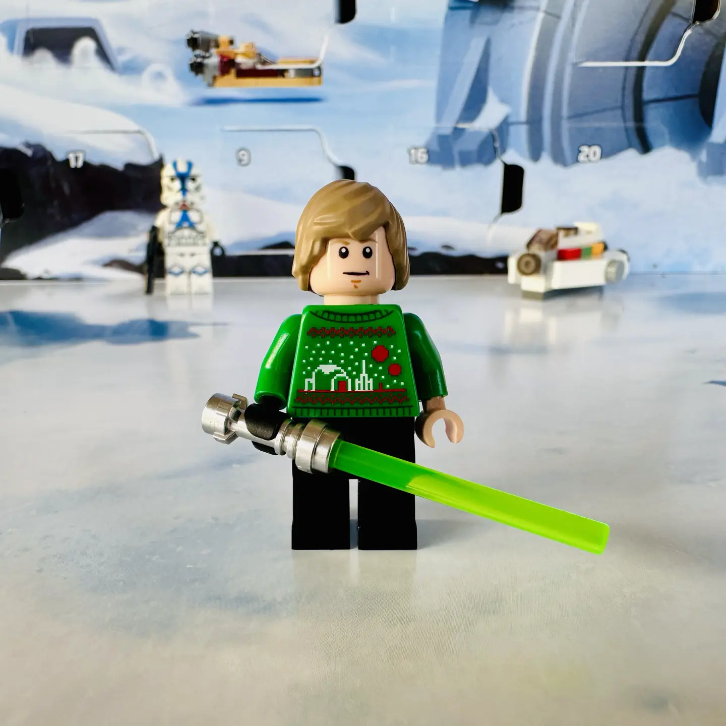 Luke Skywalker as a LEGO minifigure with a green knitted holiday sweater decorated with his homestead on Tatooine, complete with twin suns and a moisture vaporator. Luke holds his signature green light saber in one black-gloved hand.