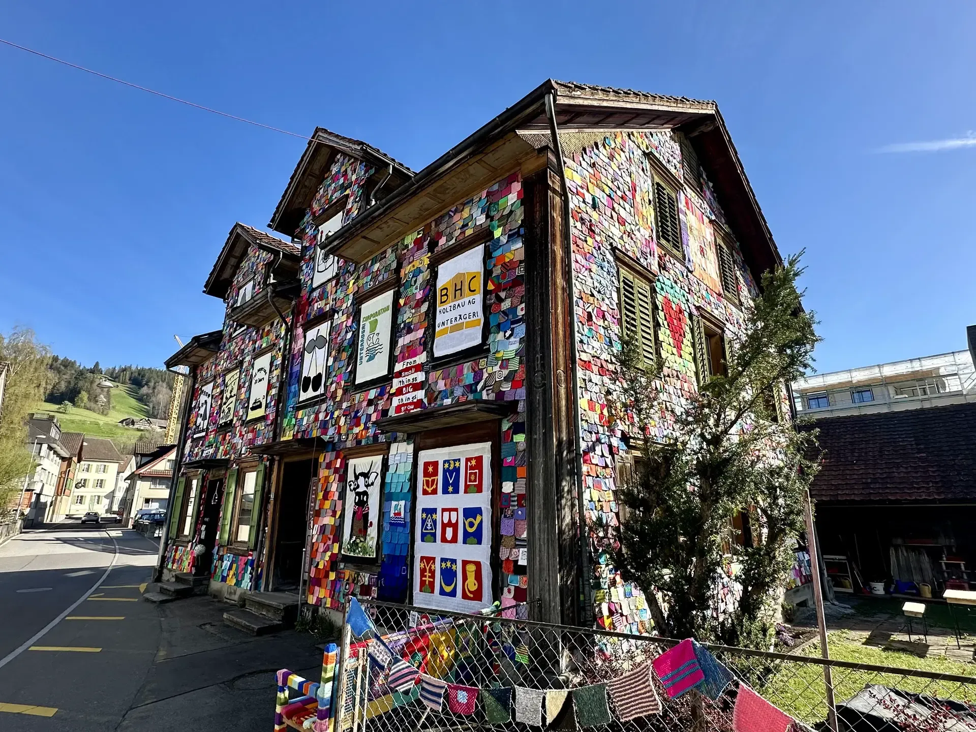 Angled view showing two sides of a historic wooden 3-story Swiss house decorated with thousands of colorful knitted yarn squares.