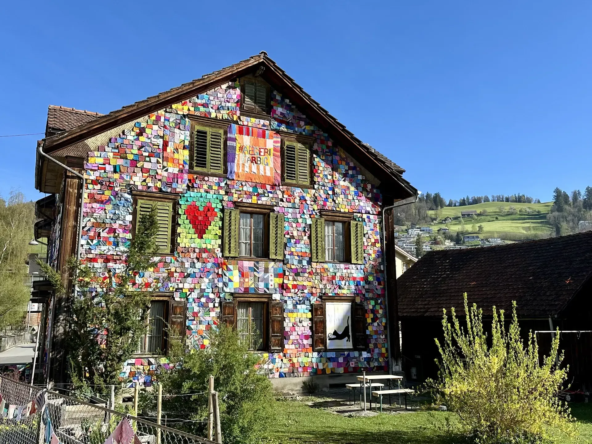 Back side and yard of a historic wooden 3-story Swiss house decorated with thousands of colorful knitted yarn squares. One large square reads "#Aegeri Farbig" and another is a pixel-art style heart.