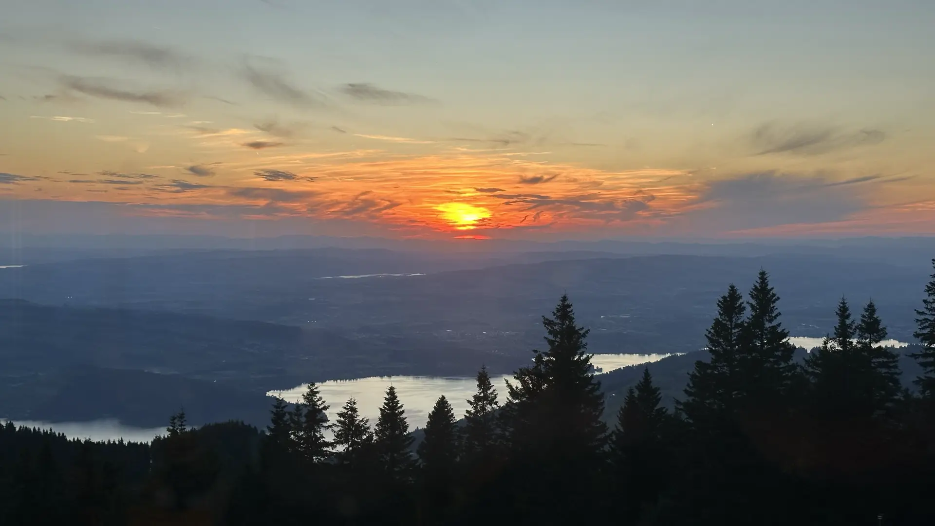 Photo of a sunset taken from the Wildspitz Berggasthaus in Switzerland. The sun is reddish orange and has nicely illuminated clouds near the horizon. A lake appears silver and pine trees form silhouettes in the foreground. Beyond the lakes are indistinct hills.