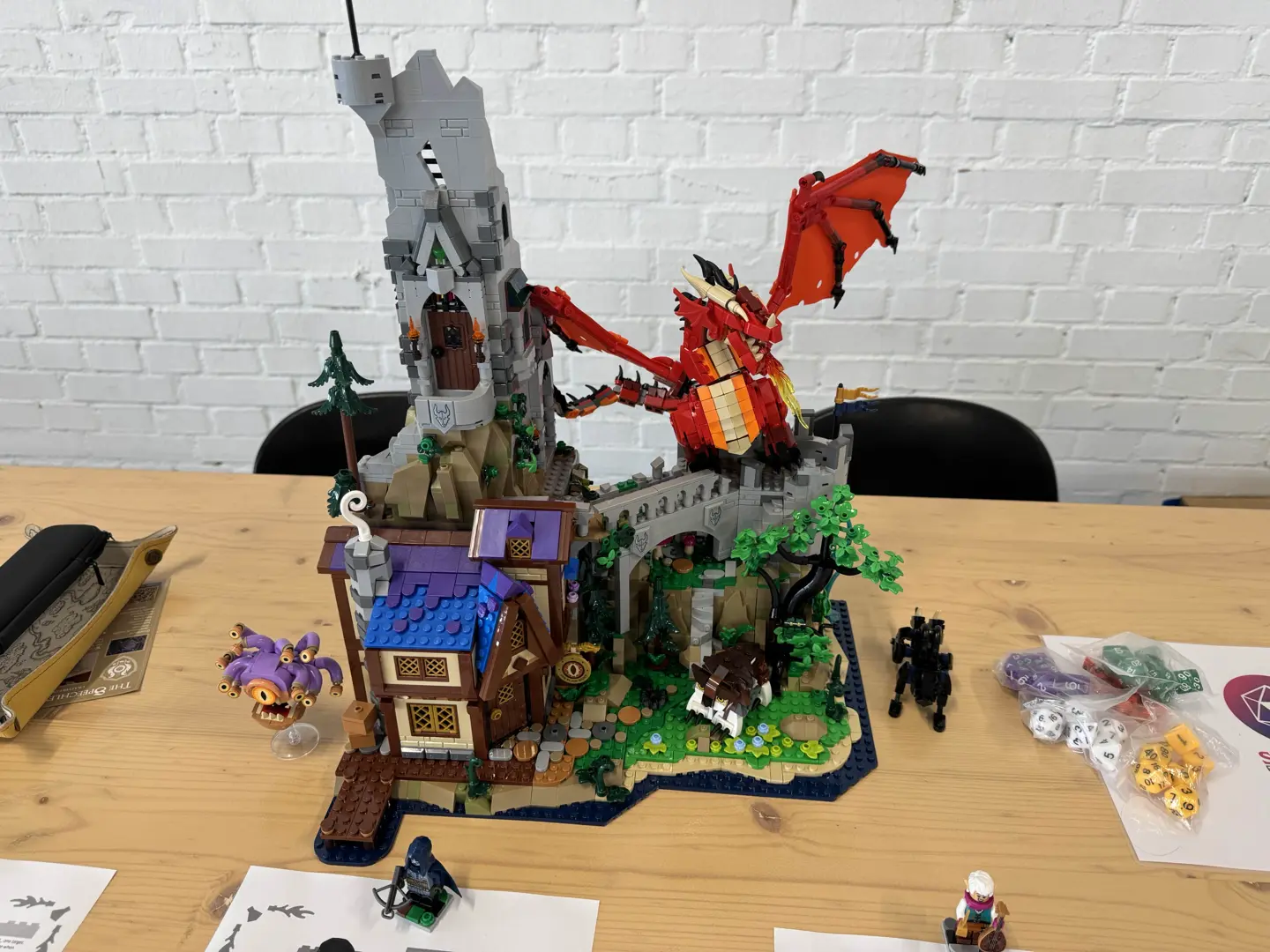 LEGO Dungeons and Dragons set 21348 topped with a red LEGO dragon on a wooden table next to some bags of dice and character sheets.