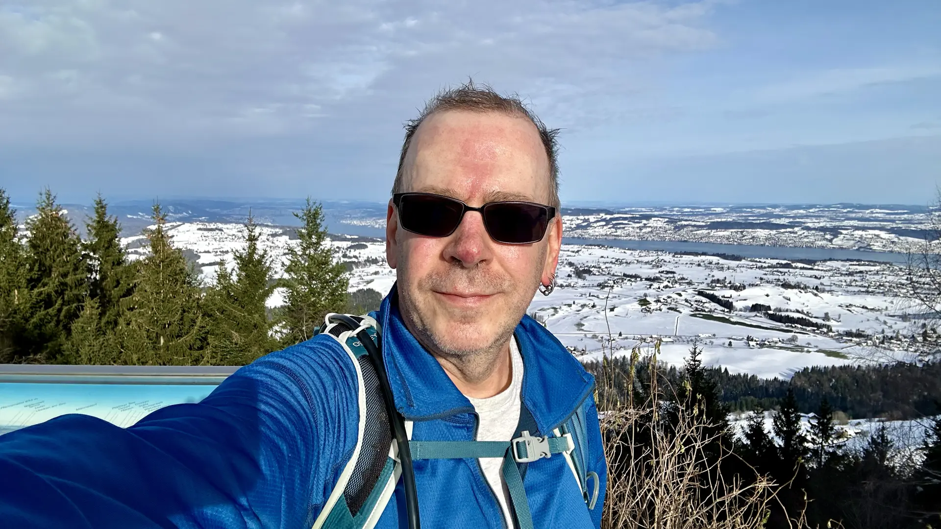 A selfie of me, a middle-aged balding white guy with tinted glasses, standing in front of a scenic view point over a winter landscape.