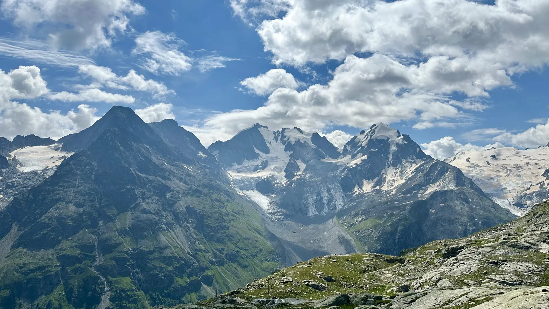 View of high mountains, some of them with glaciers. Rocks and short green grass are in the foreground. The sky is blue with multiple puffy white clouds.