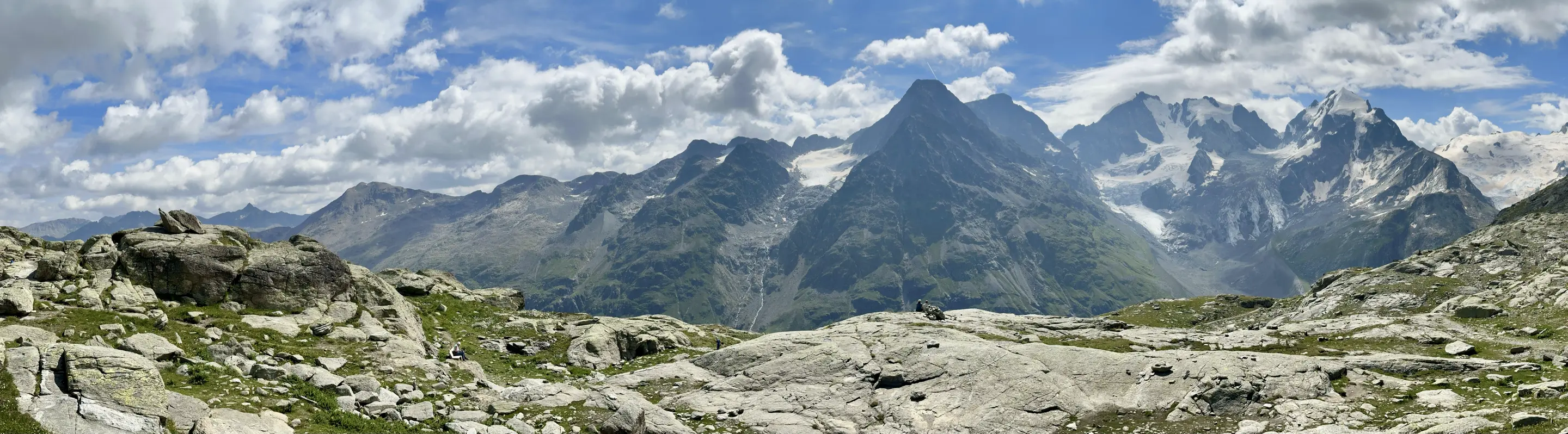 Panorama view of high mountains, some of them with glaciers. Rocks and short green grass are in the foreground. The sky is blue with multiple puffy white clouds.