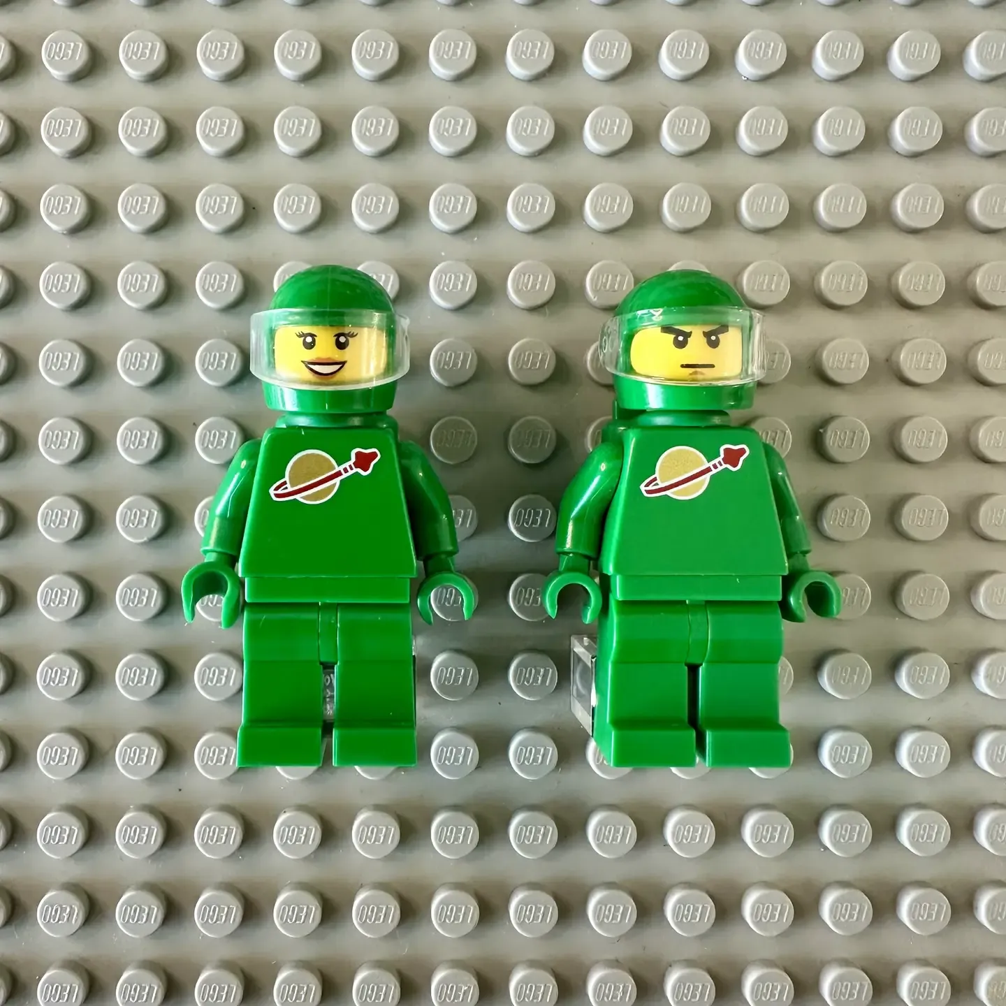 Two green astronaut LEGO minifigures from the Exo Suit set both bearing the Classic Space logo of a red ship orbiting a gold planet on their torsos. On the left is a smiling woman and on the right is a man with an angry expression.