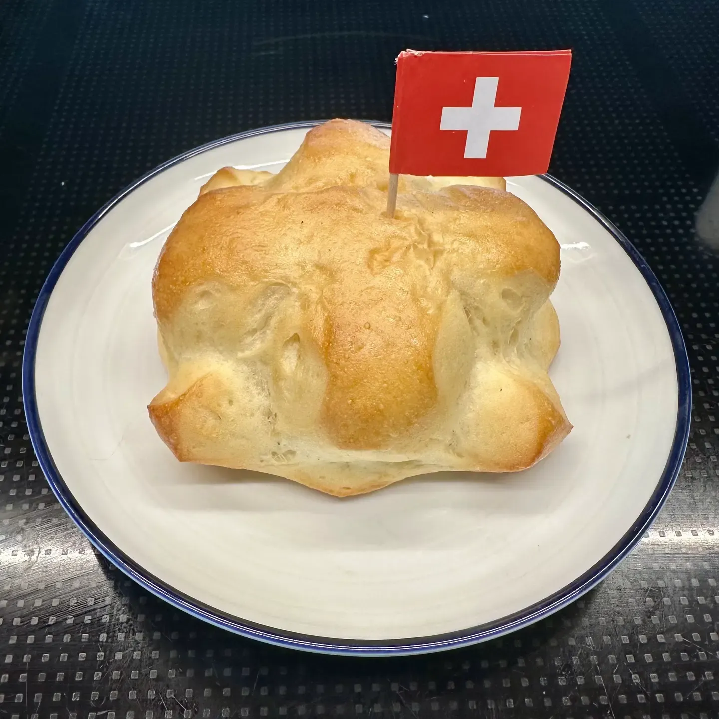 A plate holding a Swiss weggli bread roll with a cross shape on top and a tiny paper Swiss flag stuck in it.