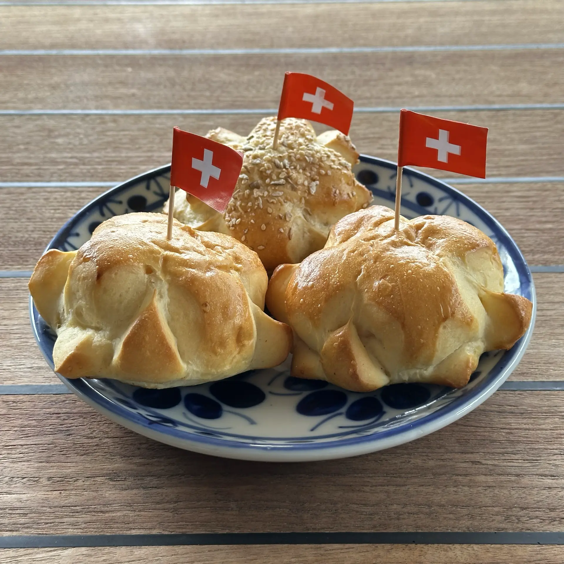 A plate holding three Swiss weggli bread rolls each with a cross shape on top and a tiny paper Swiss flag stuck in them.