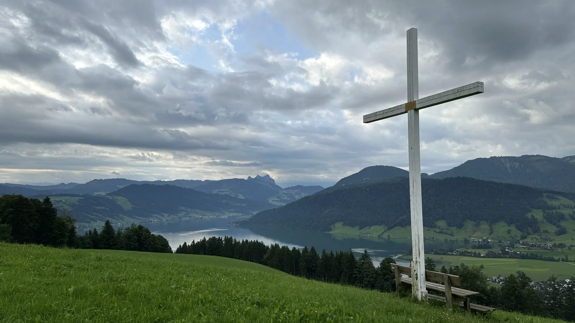 Photo view of a Swiss lake surrounded by hills on a cloudy day. A white wooden cross looms over an unpainted wooden bench in the right foreground.