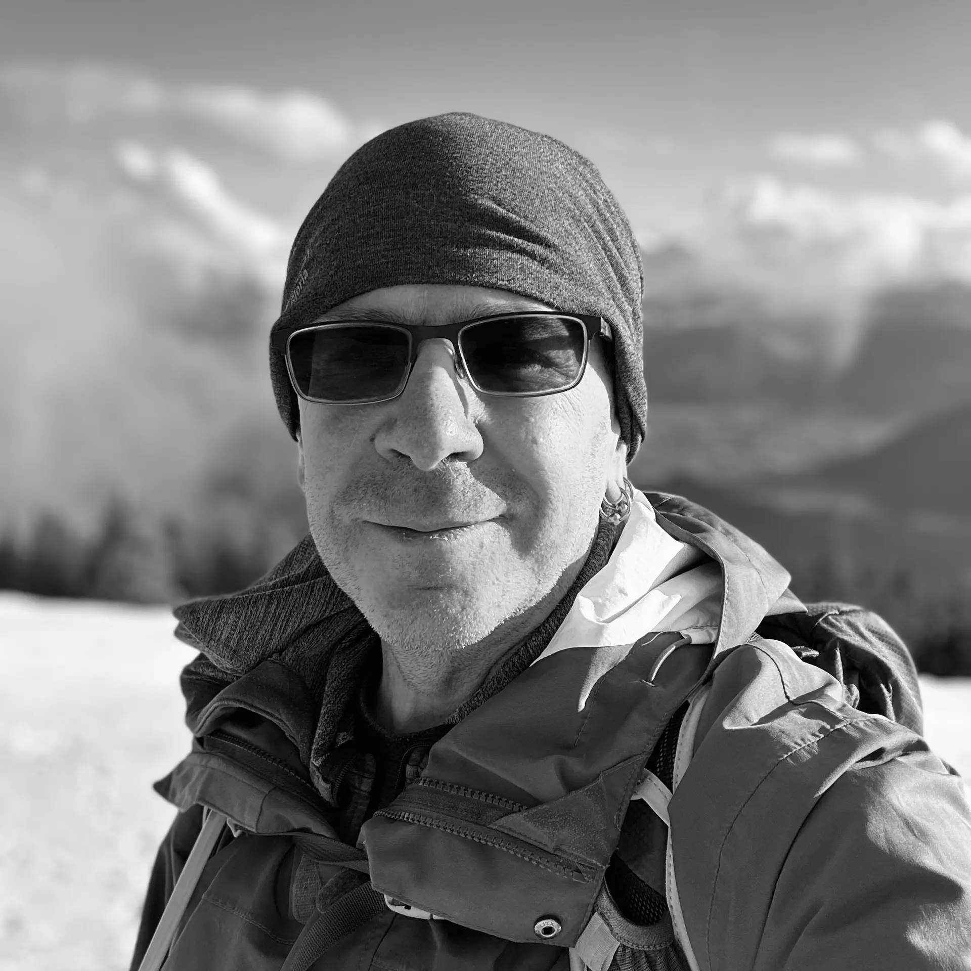 Black and white headshot photo of me, a middle-aged white man wearing sunglasses, against a blurred backdrop of mountains. Taken on top of the Wildspitz on the border between Cantons Zug and Schwyz, Switzerland.