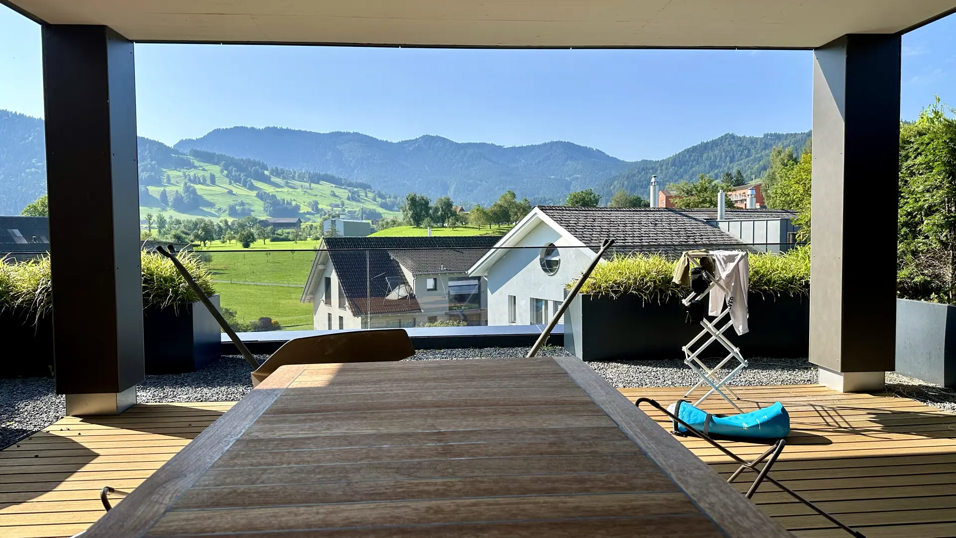 A wooden table situated on a wood-floored terrace with a view of a few multi-family houses, green pastures, and a forested mountain in the distance under a hazy light blue sky.