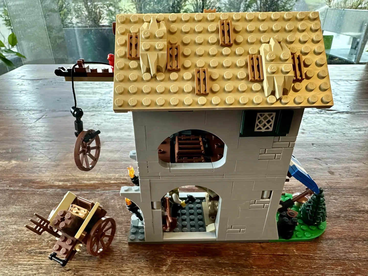 LEGO two story gray stone woodworker's shop covered with a thatched roof. A two-wheeled peasant's cart carrying a barrel and a pitchfork sits outside.