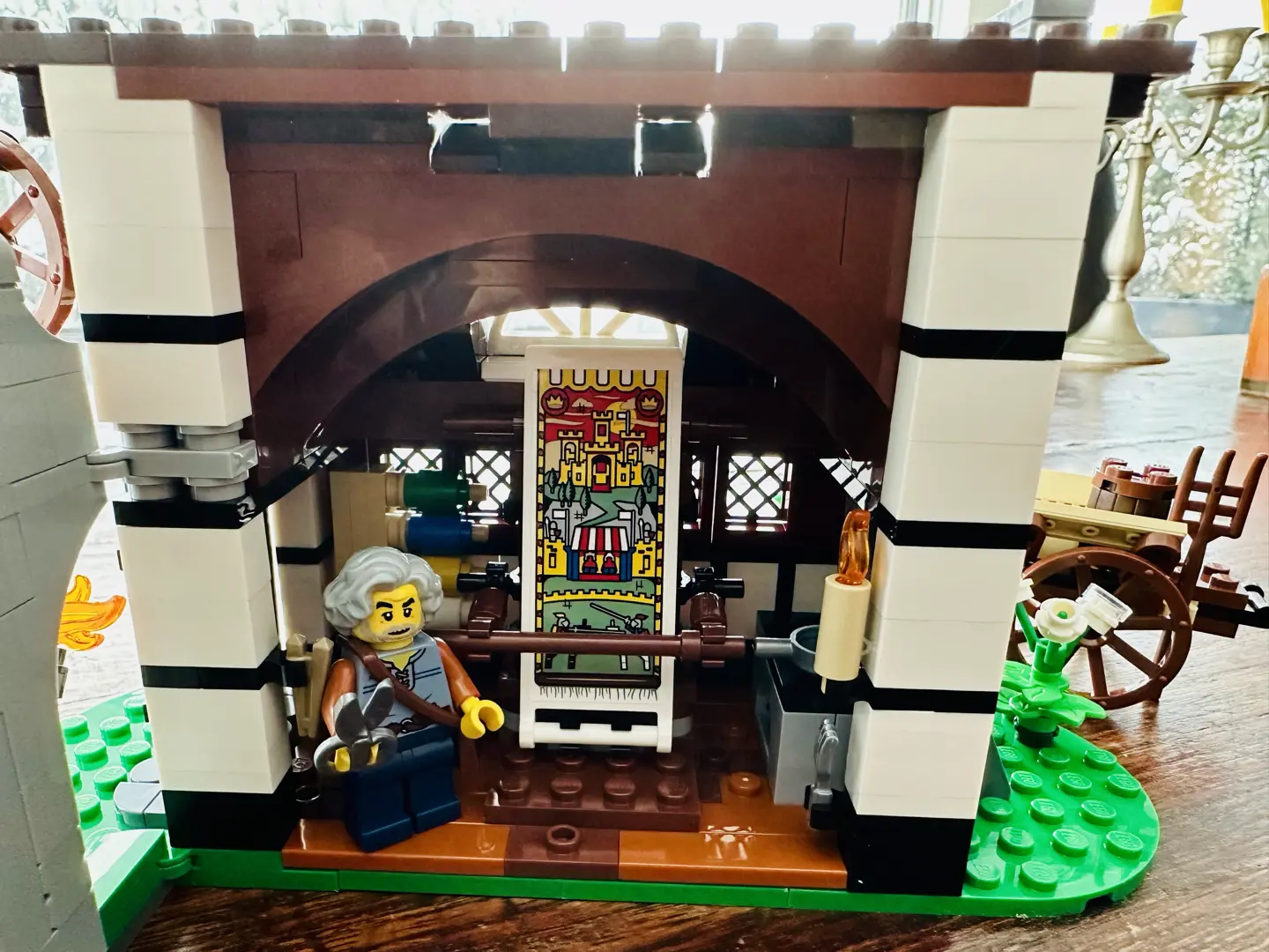 Interior of LEGO farmhouse featuring a loom depicting a scene with a castle and knights jousting. A LEGO minifig of an older man with wavy gray hair holds a pair of shears.