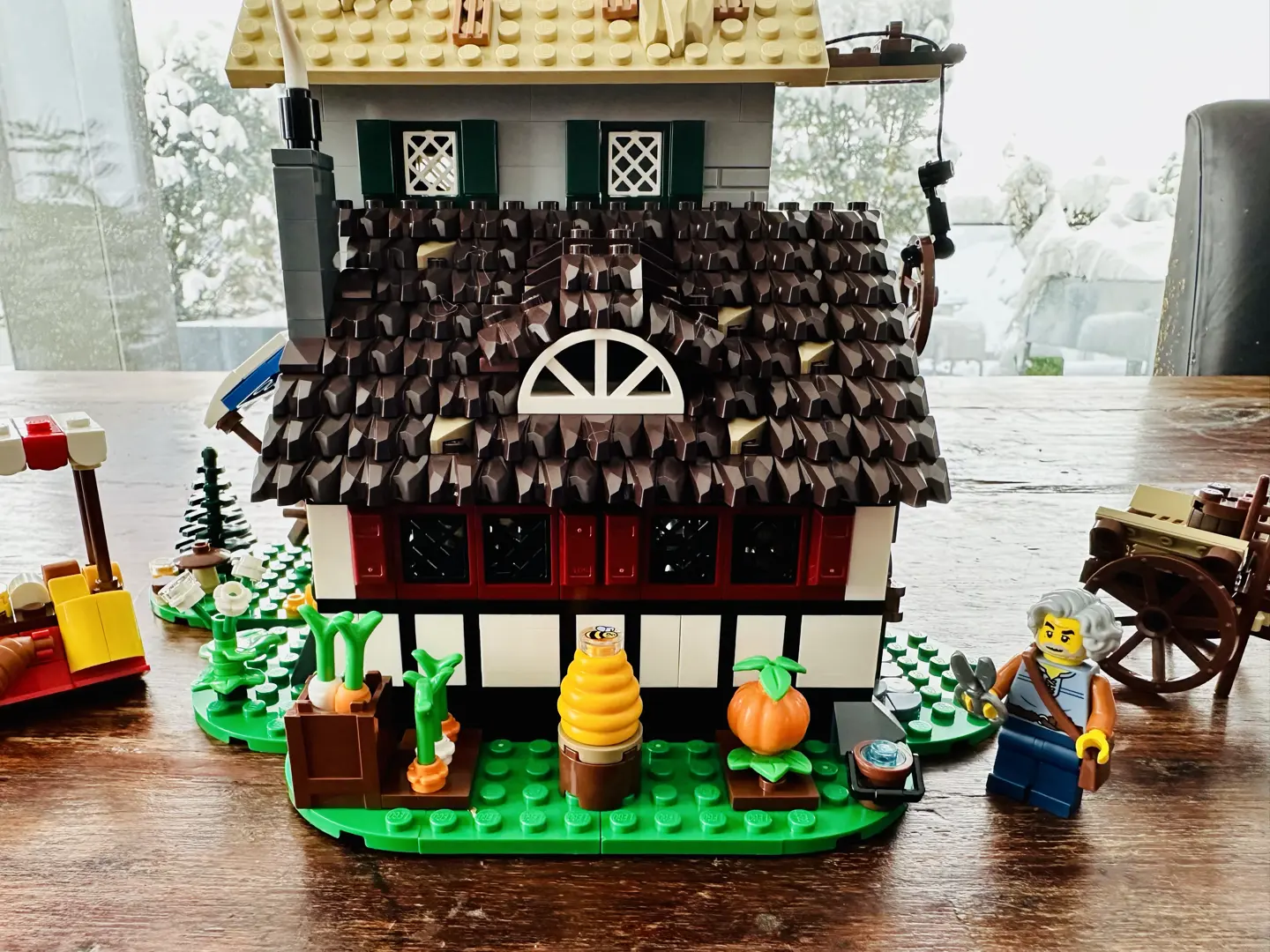 Exterior of LEGO darkly thatched farmhouse featuring a garden with carrots, pumpkins, beehive, and a pail of water. A LEGO minifig of an older man with wavy gray hair holds a pair of shears.