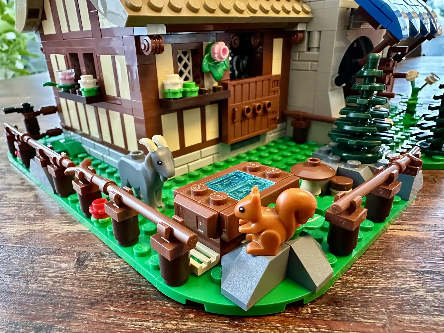 Garden of LEGO cheesemaker's shop with gray goat, squirrel, water trough, wood rail fence, flowers, and a few stones.