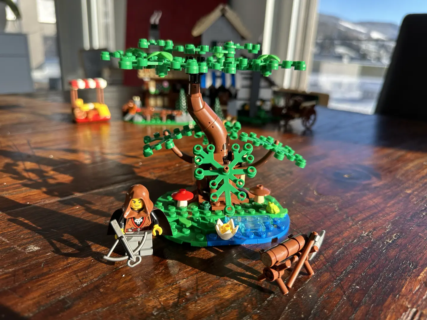 A gnarled and leafy LEGO tree by a patch of water. There are mushrooms, a lily flower, and a frog. A main wearing a brown hood and wolf-insignia tunic carries a crossbow. An axe leans against a saw horse holding a log.