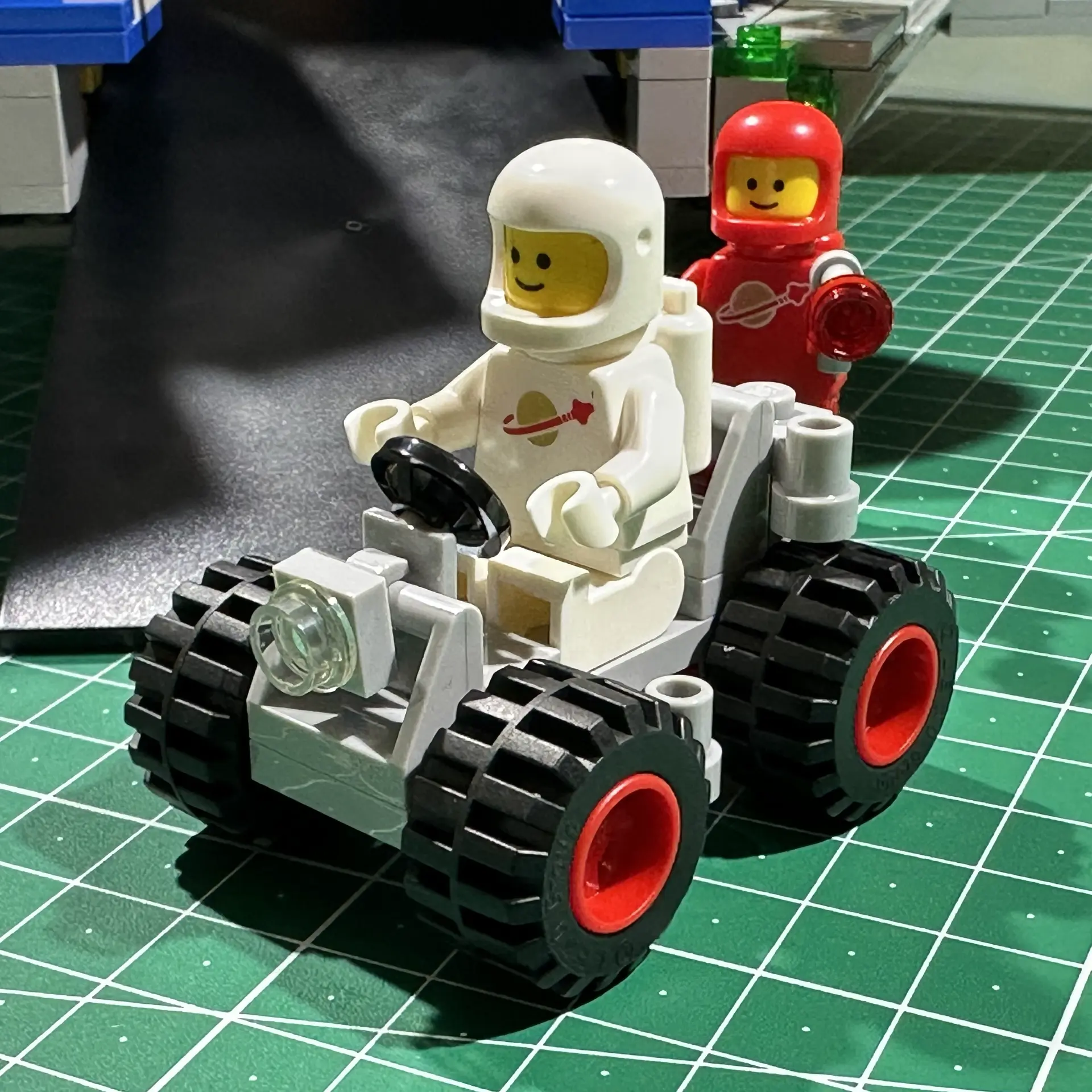 A LEGO astronaut drives a small gray rover while another astronaut looks on