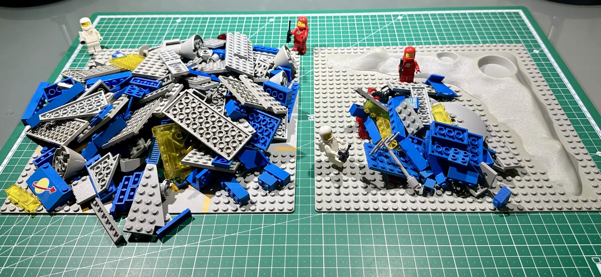 Two piles of loose vintage LEGO pieces (predominately gray and blue) on two baseplates with four astronaut minifigures posed around them. These are the pieces for the original Galaxy Explorer, set 497 from 1979