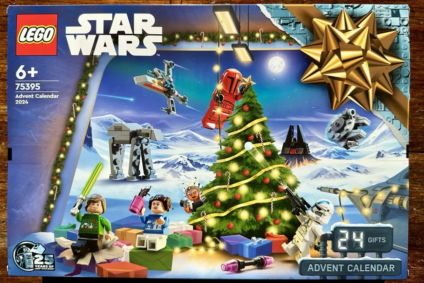 Box for LEGO Star Wars Advent Calendar 2024. It depicts a big golden bow, a Christmas tree, LEGO minifigs of Luke Skywalker, Princess Leia, Ahsoka Tano, and two armored troopers plus multiple tiny LEGO space vehicles.