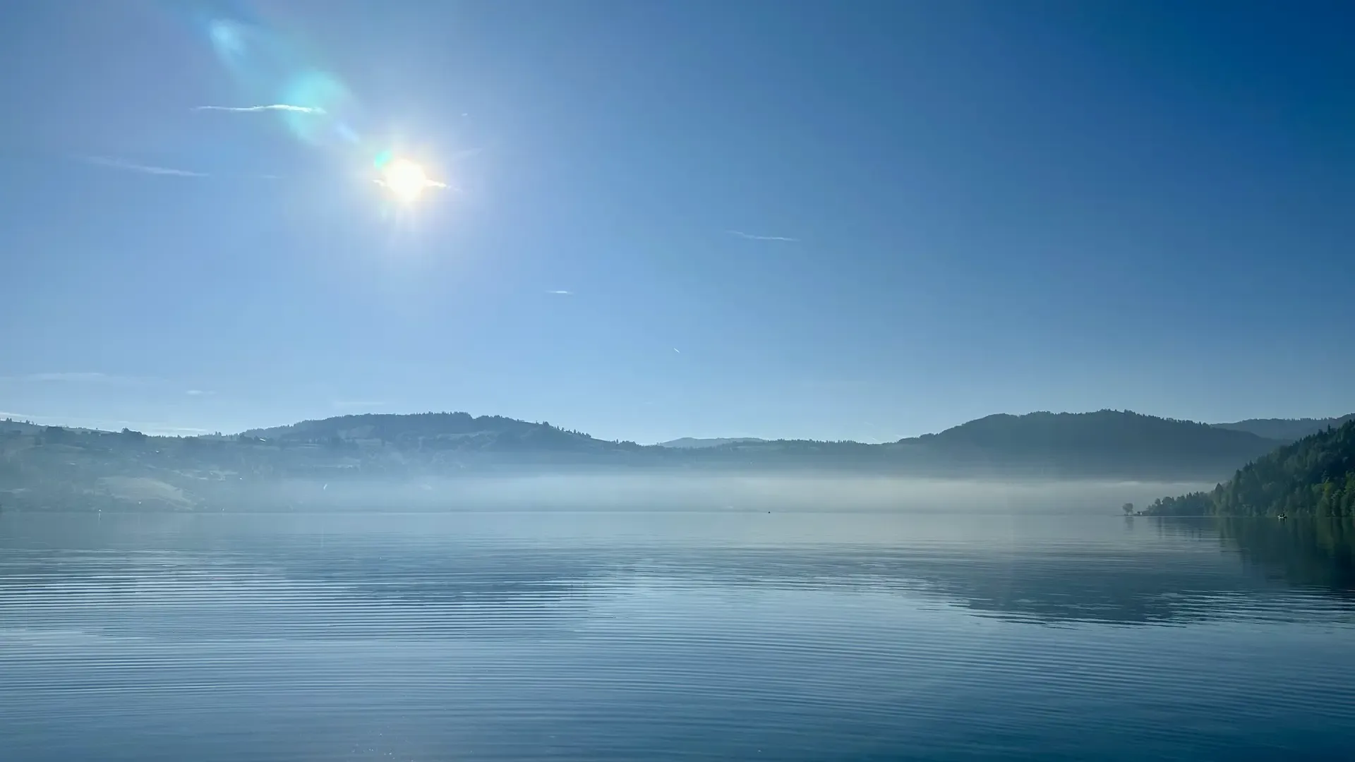 Morning view of a calm lake under a cloudless blue sky. A bit of mist (or perhaps just a low-lying cloud) hangs over the surface of the lake in the distance.