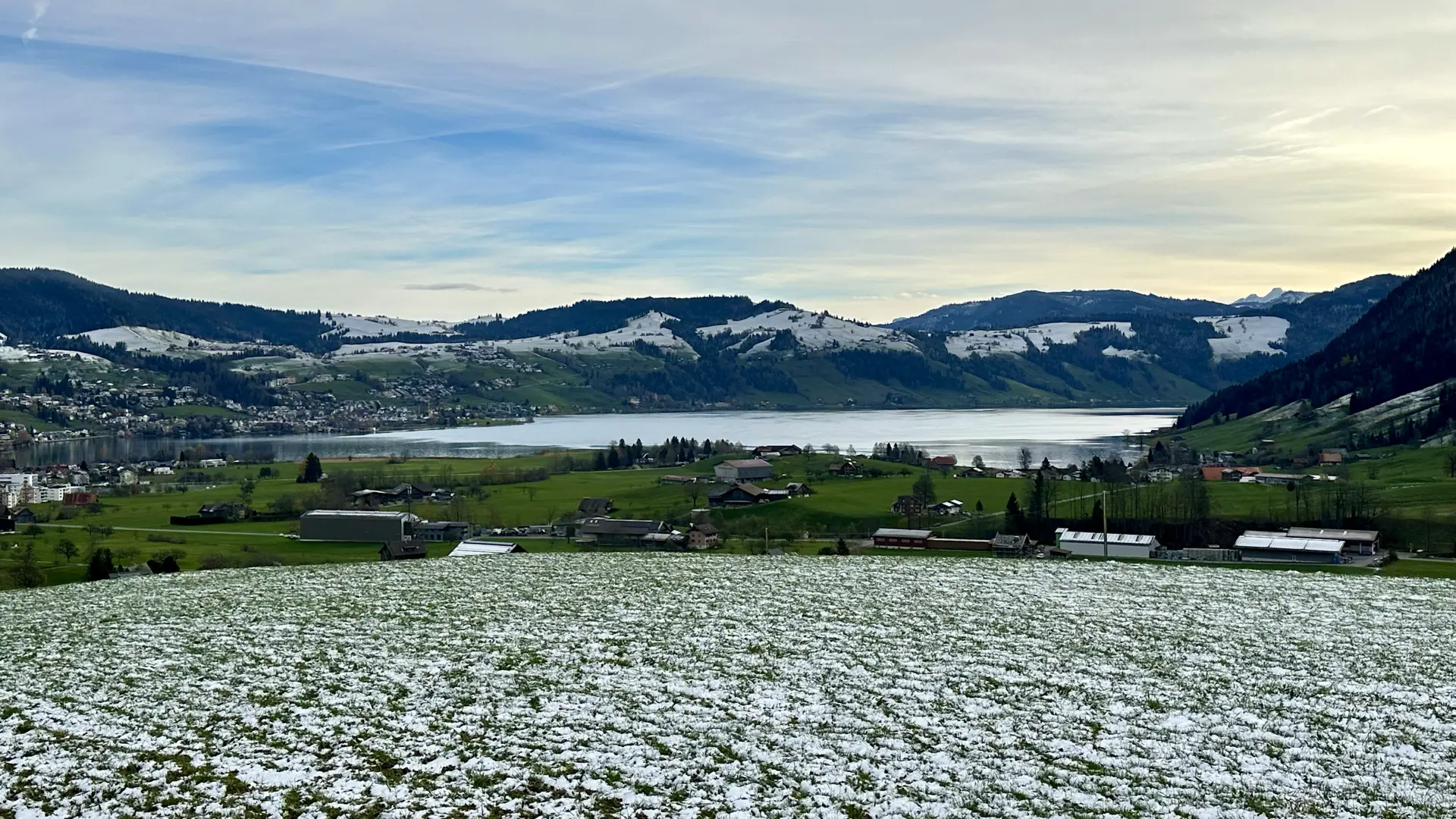 Landscape view over partially snow-covered farm fields towards a lake. Beyond the lake are partially forested and partially snow-covered hills under a cloudy sky with chemtrails and some blue showing.