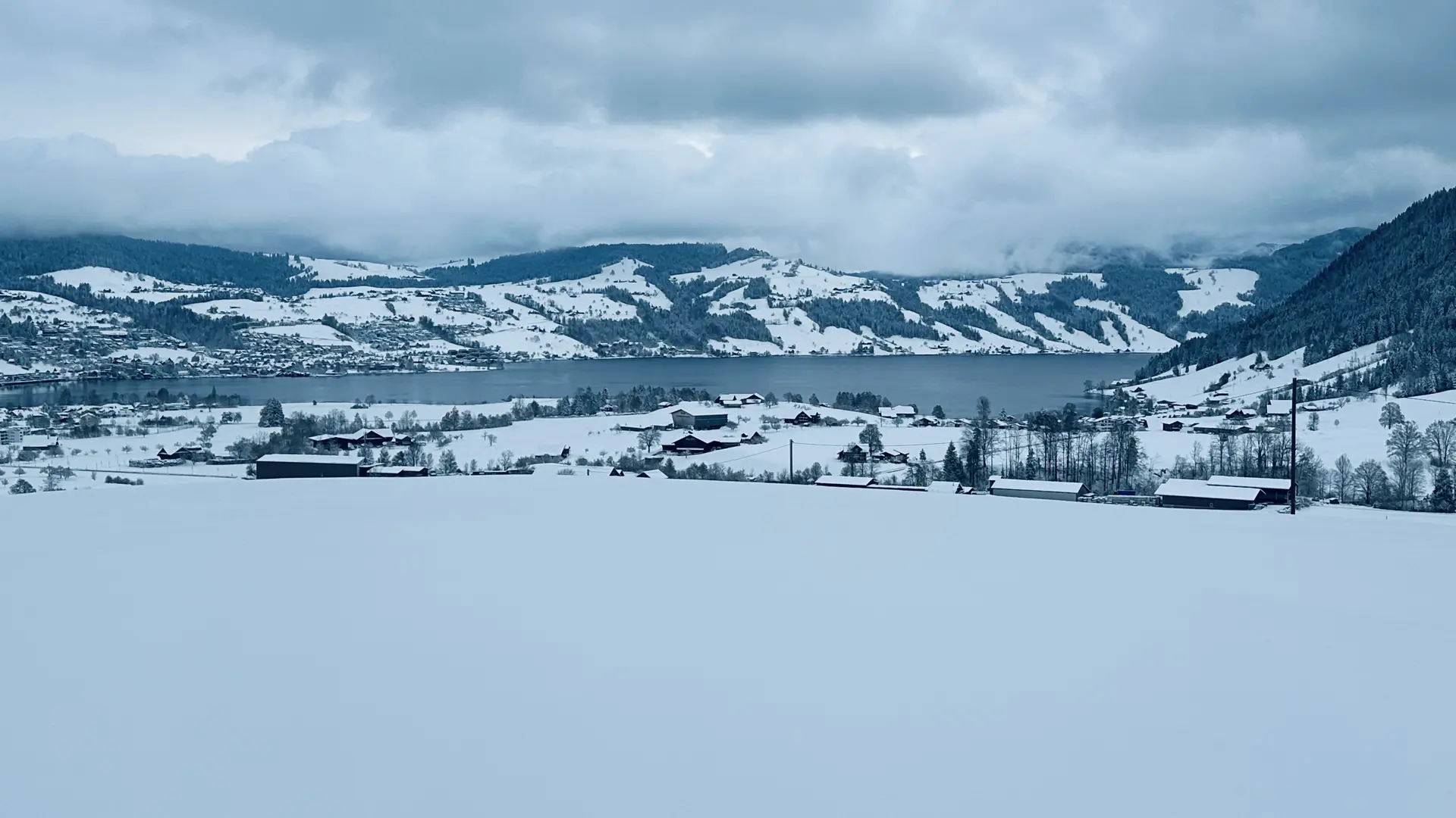 Landscape view over snow-covered farm fields towards a lake. Beyond the lake are partially forested snow-covered hills under an overcast sky with dramatic clouds.