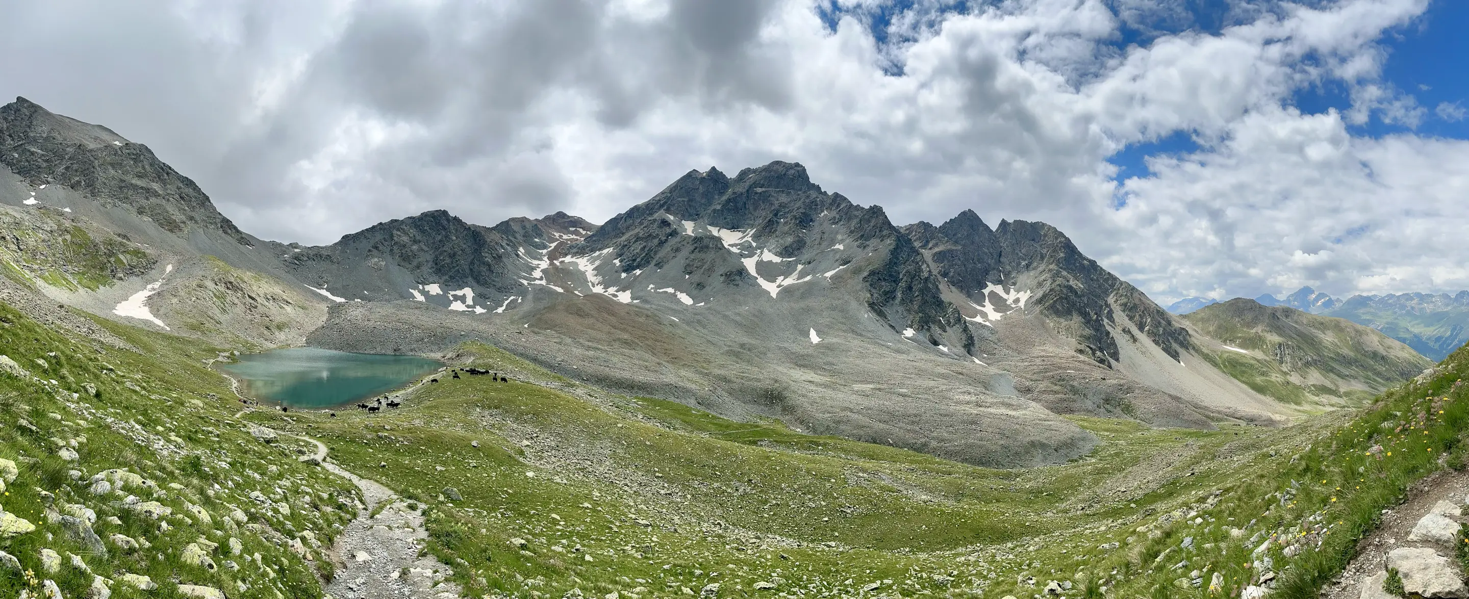 Panorama of high alpine terrain with a small blue green lake on the left, dark gray craggy mountains dotted with a bit of old snow in the distance, and green heather dotted with white stone in the foreground.