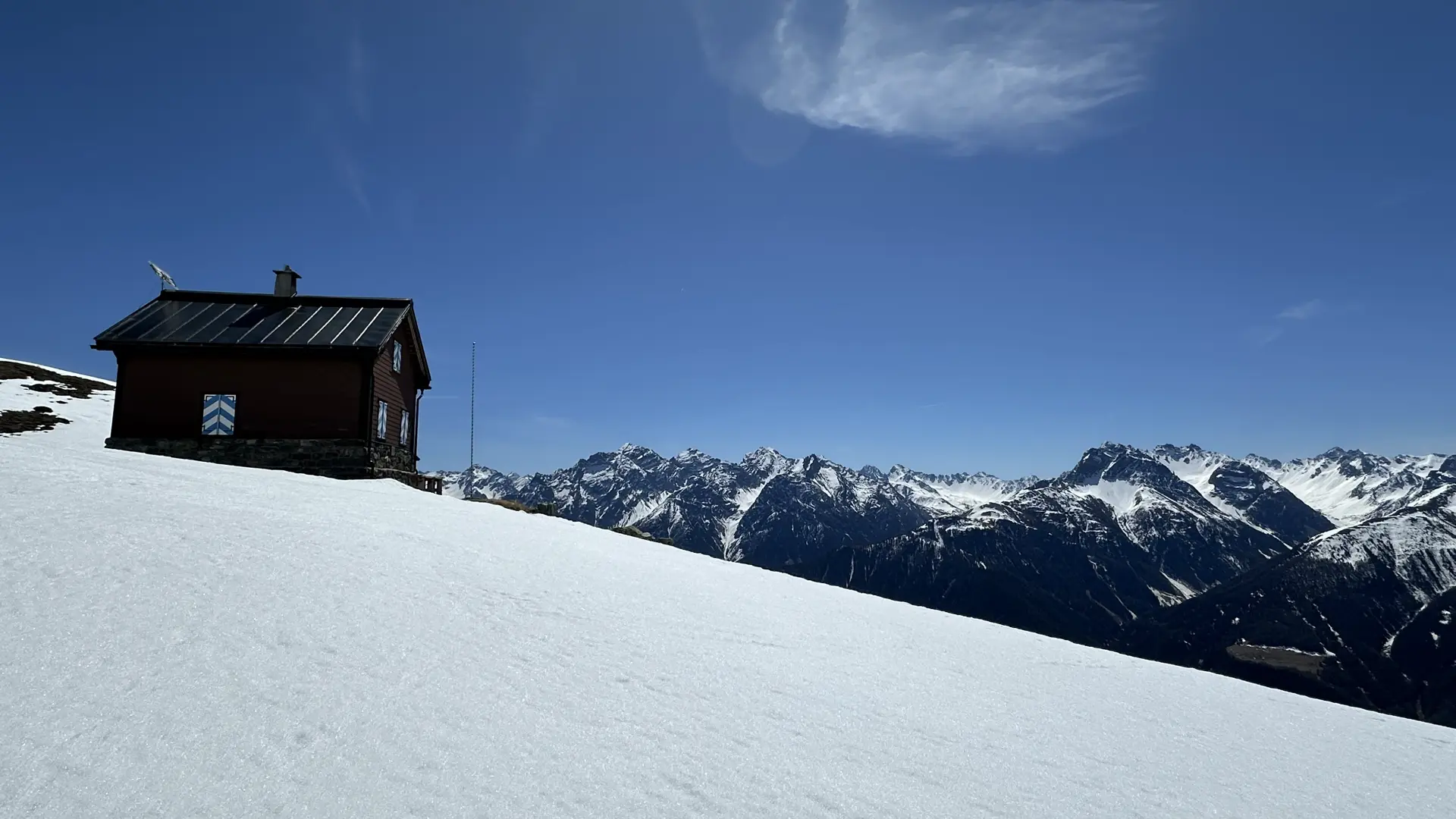 Small brown hut with blue and white chevron shutters perched on a snowy hill overlooking the Alps under a blue sky.
