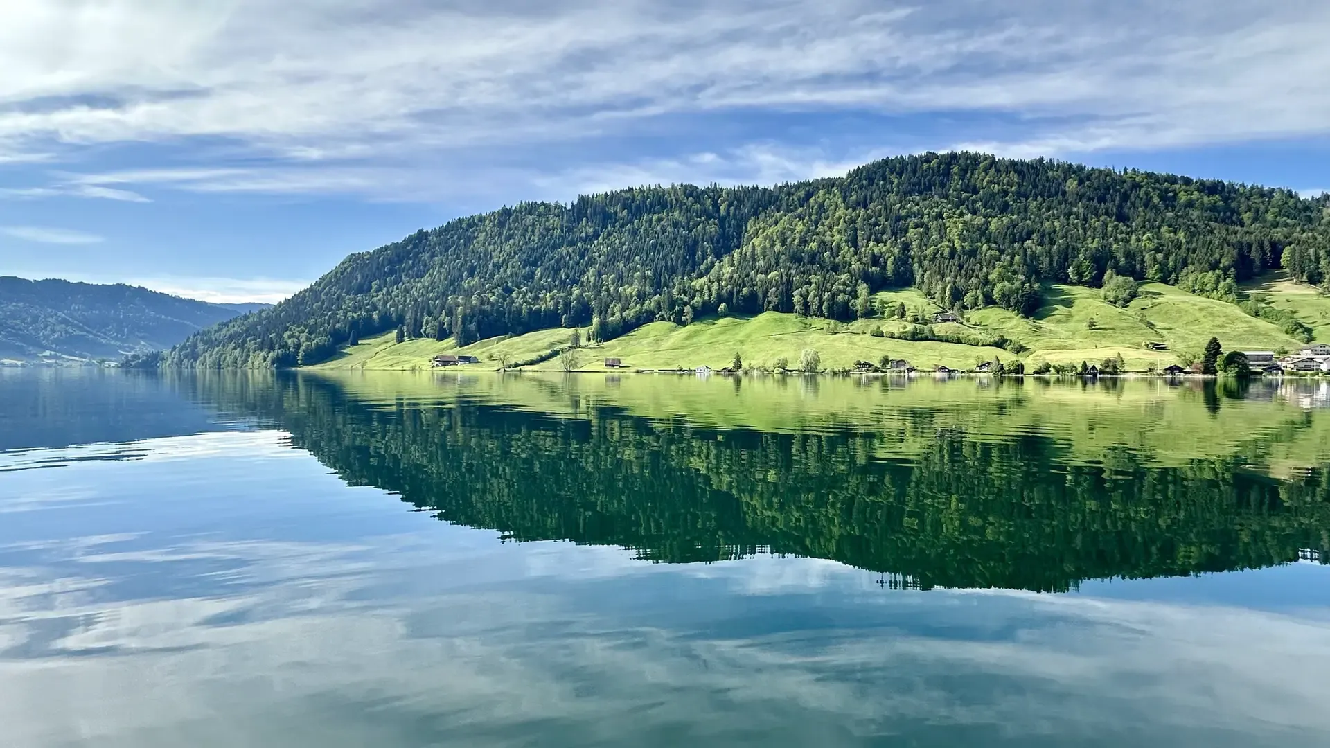 Green pasture and forested hills under a blue sky with white clouds reflected in a calm lake.