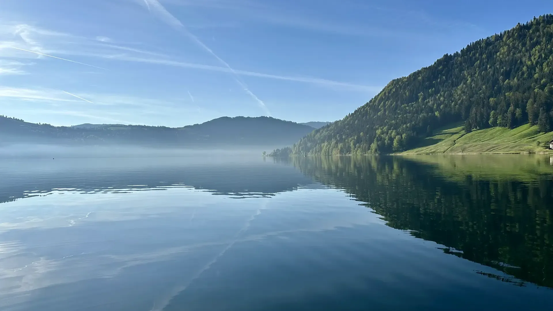 View of a calm lake under a blue almost cloudless sky showing a few contrails. Green forested hills are on the right side, while on the left mist hovers over the surface of the lake in the distance. Everything is reflected in the surface of the lake.