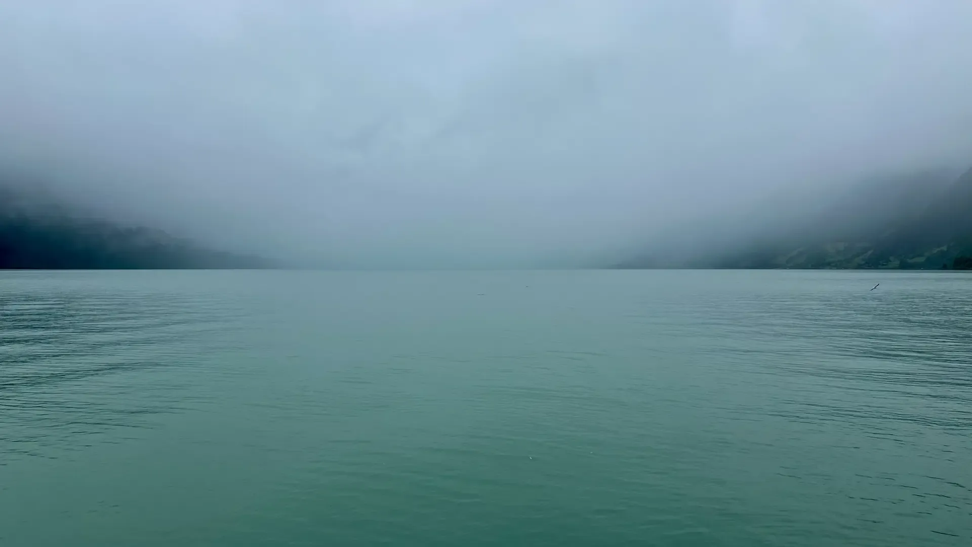 View of a lake on a very misty day. The water is blue-green.