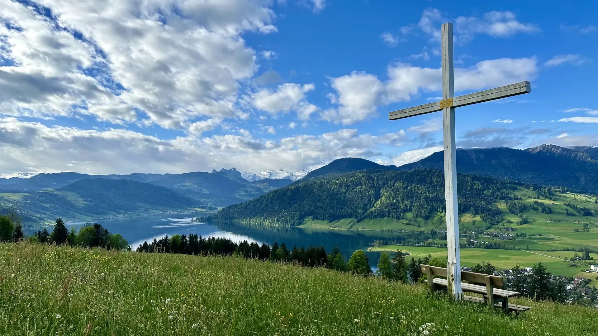 View of a lake on a lovely day. Green pasture dotted with yellow flowers is in the foreground together with a park bench under a large white cross. The sky is blue with many puffy white clouds.