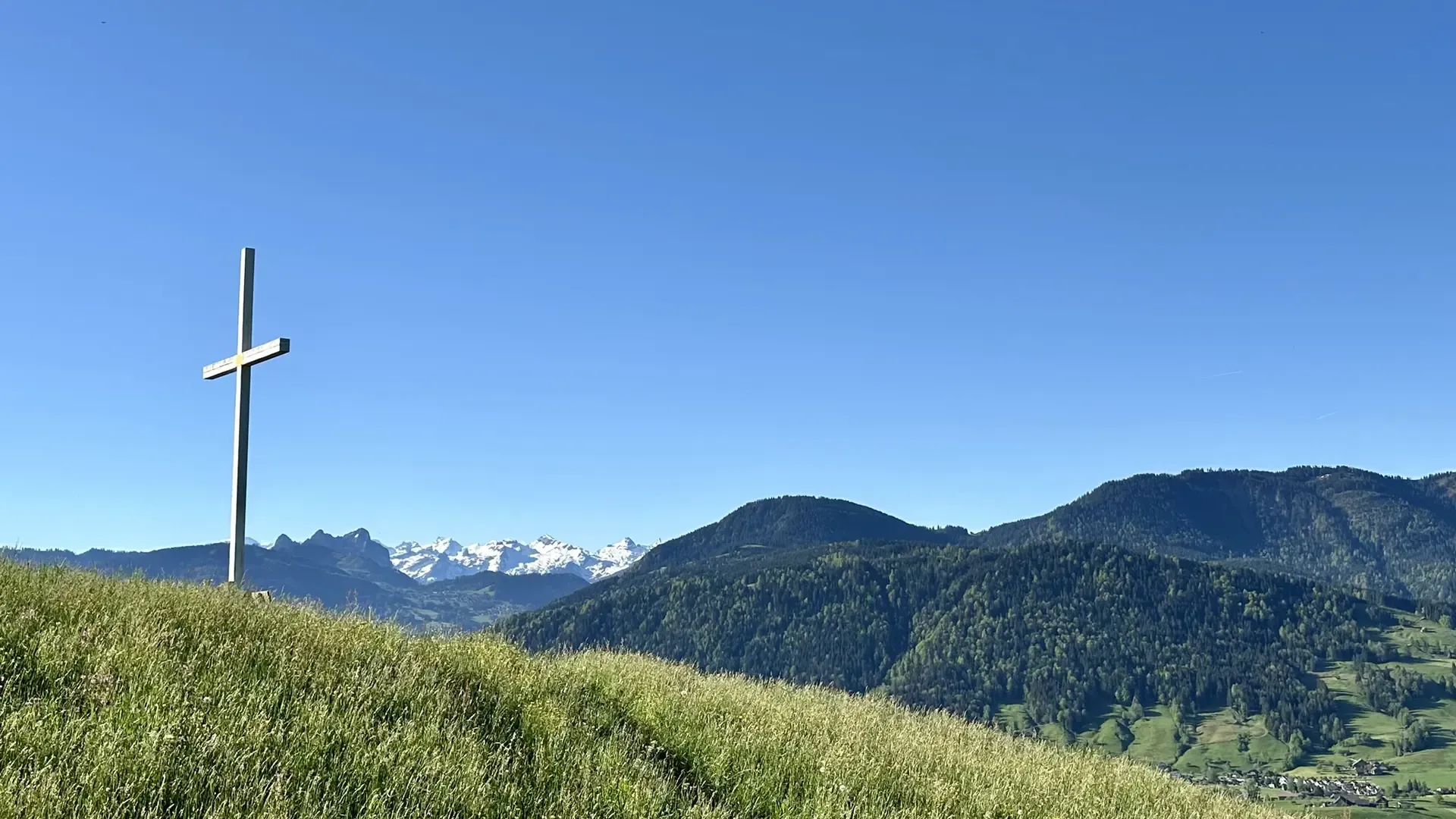 View of light green pasture and dark green forested hills on a sunny day. A large white cross is silhouetted against a cloudless blue sky. A few snow-covered mountains are visible in the far distance.