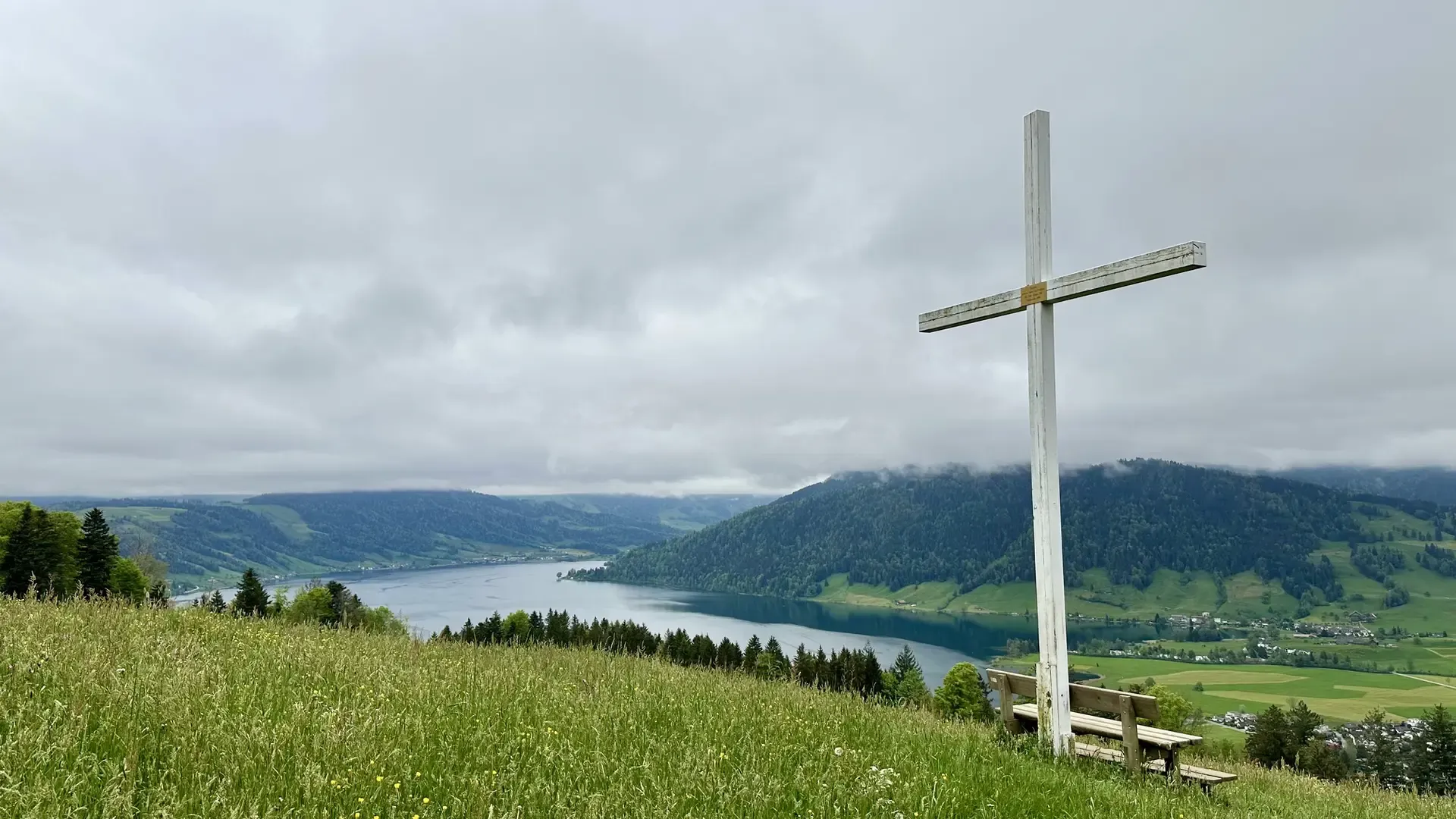 View of a lake on an overcast day. Green pasture dotted with yellow flowers is in the foreground together with a park bench under a large white cross. The sky is completely covered in clouds.