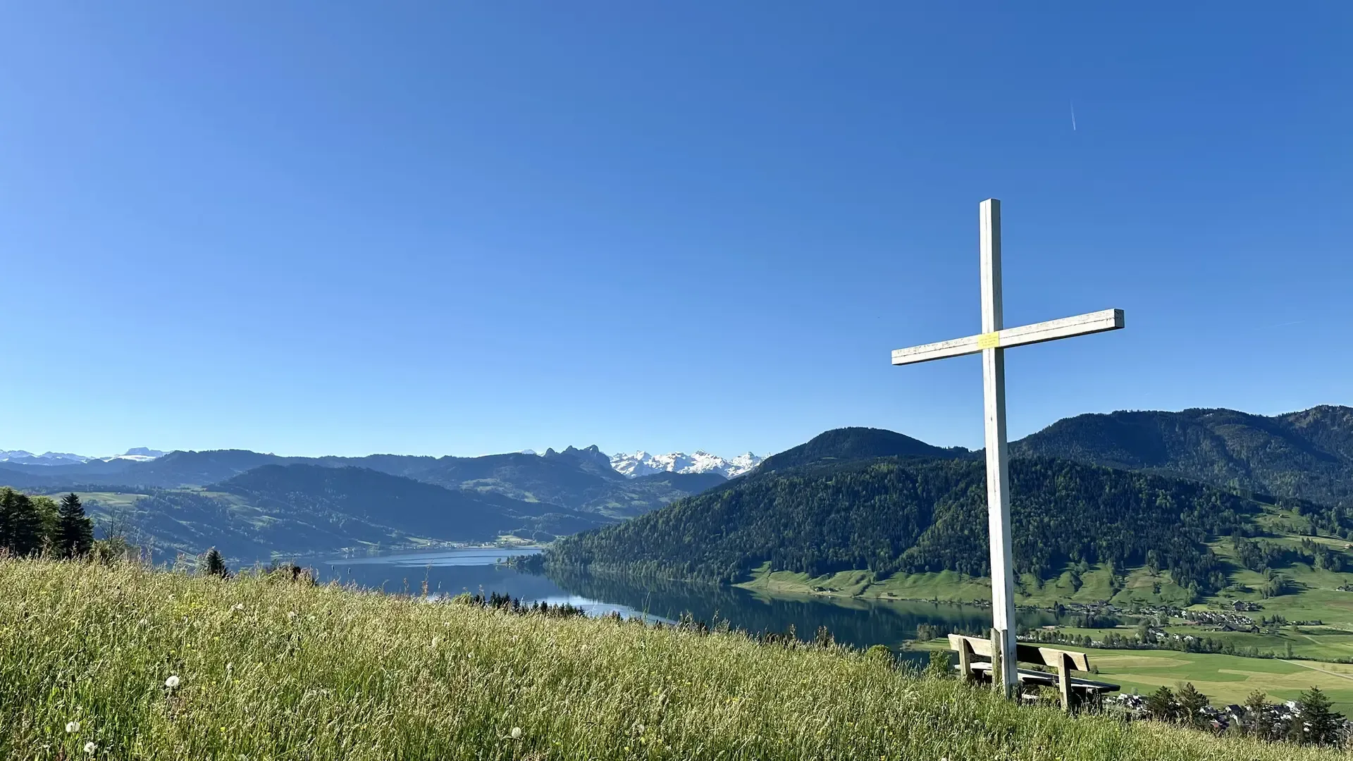 View of a lake on a sunny day. Green pasture dotted with yellow flowers is in the foreground together with a park bench under a large white cross. The sky is cloudless and blue.